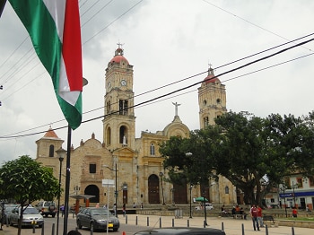 Mujer con sombrero de paja y gafas de sol sostiene un helado rosa en cono frente a letras de colores y una iglesia de dos torres