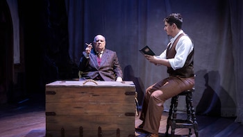 Dos hombres con vestimenta de época en un escenario teatral, uno sentado junto a un baúl y el otro en un taburete con un libro en las manos