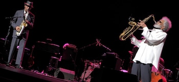 Dos saxofonistas actúan en un escenario oscuro con luces púrpuras. El de la derecha, Sonny Rollins, viste camisa blanca y el de la izquierda, traje gris