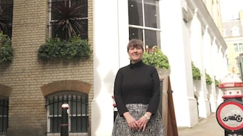 Mujer de cabello castaño corto, suéter negro y falda a cuadros, de pie frente a un edificio de ladrillo con ventanas y jardineras, sonriendo a la cámara