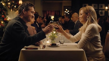 Un hombre y una mujer sonrientes brindan con copas de vino tinto en una mesa de restaurante iluminada, con un árbol de Navidad visible al fondo