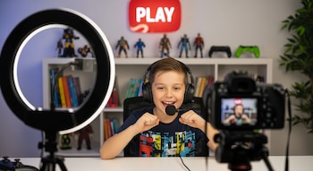 Niño sonriente con auriculares y micrófono grabando un video, frente a una cámara en trípode y un aro de luz. Fondo con estantes de libros y juguetes.