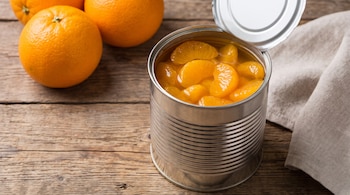 Una lata de conserva abierta con gajos de naranja en almíbar, tres naranjas enteras, una superficie de madera y un paño de tela.