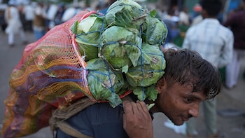 Primer plano de un hombre que lleva en su espalda una gran bolsa de malla roja llena de repollos verdes y otras verduras