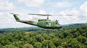 Un helicóptero verde con la inscripción AN-146 Panamá vuela sobre la copa de los árboles bajo un cielo parcialmente nublado.