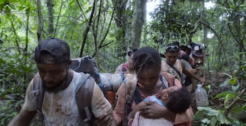 Grupo de personas camina por un sendero estrecho en la selva. Un hombre con gorra y mochila, una mujer lleva un bebé. Otros individuos con mochilas. Vegetación densa