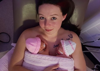 Mujer acostada sosteniendo a dos bebés prematuras envueltas en una manta rosa, ambas con gorros de lana y tubos médicos en sus rostros