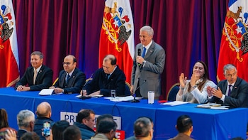 Un hombre de pie con micrófono habla ante un panel de funcionarios sentados en una mesa azul. Hay banderas chilenas y cortinas rojas al fondo, y el público es visible