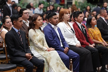 Fila de seis personas sentadas en sillas de madera con cojines. Hombres con traje, mujeres con vestidos o blusas formales. Fondo borroso con más gente