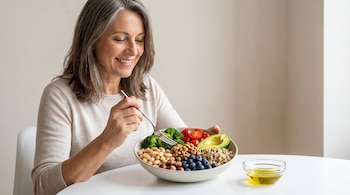 Mujer sonriente de 51 años en mesa blanca, comiendo un bol con brócoli, tomates, aguacate, frijoles, nueces, arándanos y quinua. Hay aceite de oliva.