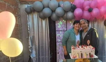 Casal, homem e mulher, sorrindo enquanto se abraçam em frente a um bolo de aniversário decorado com morangos, com balões cinza e rosa ao fundo.