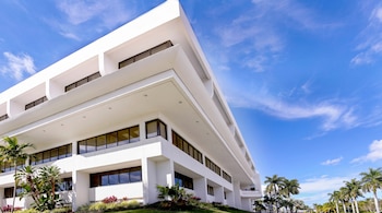 Edificio blanco de varios pisos con ventanas oscuras, palmeras verdes y otra vegetación, bajo un cielo azul con nubes dispersas.