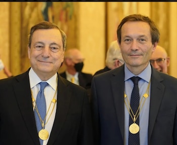 Primer plano de dos hombres con trajes oscuros, camisas blancas y corbatas, luciendo grandes medallas doradas alrededor del cuello, en un entorno formal