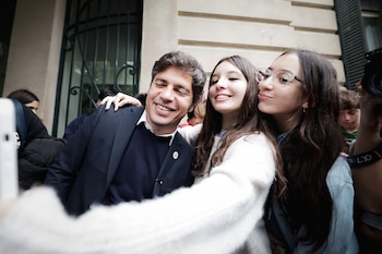 Axel Kicillof, un hombre de cabello oscuro y traje azul marino, sonríe en una selfie con dos jóvenes mujeres, una con suéter blanco y otra con gafas