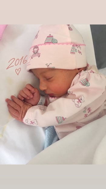 Una bebé recién nacida duerme vestida con pijama y gorro rosados, con la inscripción de 2016 y un corazón dibujado en la almohada