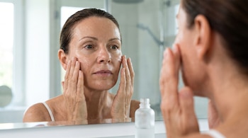Mujer adulta aplicando sérum transparente en su rostro frente a un espejo, con gotas visibles en su mejilla, y un frasco de sérum en primer plano.