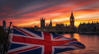 Una bandera del Reino Unido ondea en primer plano. Detrás, el Palacio de Westminster, el Big Ben y el río Támesis al atardecer con un cielo naranja intenso.