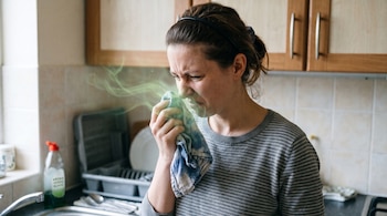 Una mujer sostiene un paño cerca de su rostro del que sale humo verde, junto a un fregadero en una cocina.