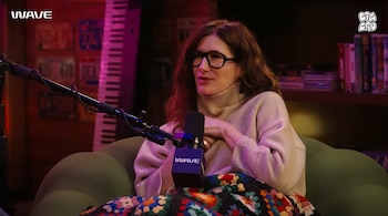 Una mujer con gafas y suéter de cuello alto beige habla a un micrófono negro con logo WAVE, sentada en un sofá verde con una manta de ganchillo colorida