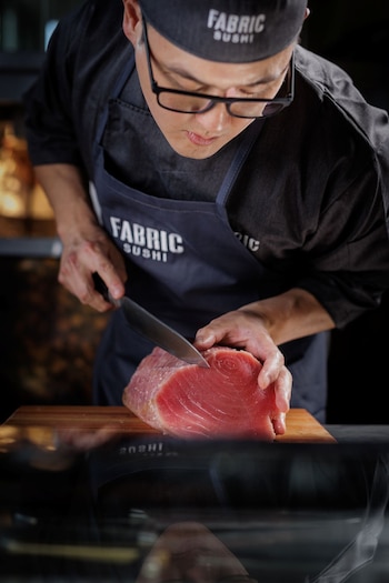 Un cocinero con gafas, gorro y delantal corta una pieza grande de atún rojo sobre una tabla de madera con un cuchillo