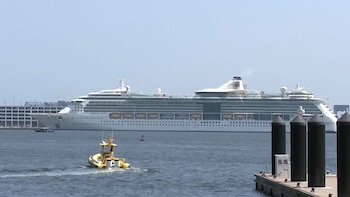Gran crucero blanco en el agua, barco amarillo pequeño con estela, muelle oscuro con postes, edificio en el fondo, cielo azul despejado