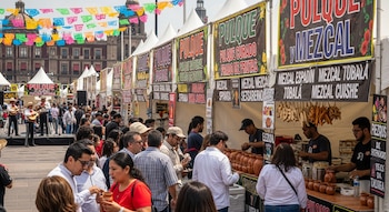 Una multitud de personas camina entre puestos de venta de pulque y mezcal bajo banderines coloridos, con mariachis tocando en un escenario a la izquierda.