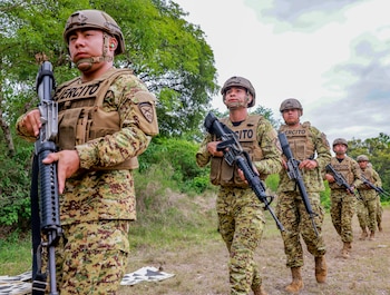 Cinco soldados con uniformes de camuflaje, cascos y chalecos tácticos caminan en fila por un campo de hierba, con árboles frondosos al fondo y cielo nublado. Todos llevan rifles