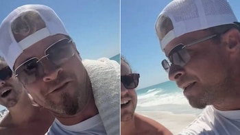 Primer plano de Brian Littrell y Alan Ritchson en la playa. Littrell lleva gorra blanca y gafas de sol, Ritchson sonríe detrás. Fondo de mar azul y arena blanca