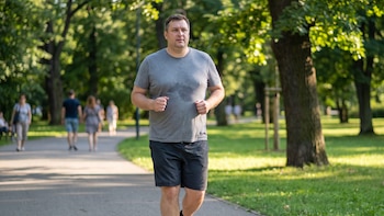 Un hombre con camiseta gris sudada y pantalón corto corre por un camino asfaltado en un parque rodeado de árboles.