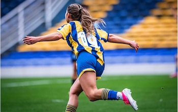 Vista de espaldas, futbolista con camiseta azul y amarilla número 19 y pantalones cortos azules corre en campo de césped verde, gradas al fondo