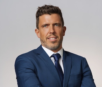 Hombre con barba corta, saco azul marino, camisa blanca y corbata azul sobre un fondo gris claro