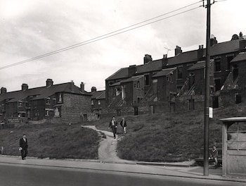 Fotografía en blanco y negro de una calle con casas adosadas de ladrillo, un camino de tierra, una acera, un poste, una parada de autobús y varias personas