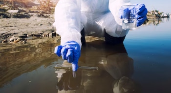 Persona con traje protector blanco y guantes azules arrodillada en agua. Sostiene un tubo de ensayo sumergido y un vaso de precipitados con líquido claro