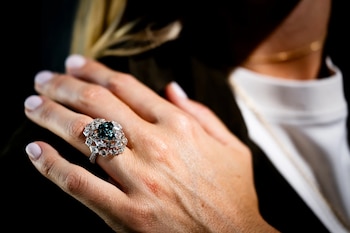 Primer plano de una mano con un anillo que lleva un gran diamante central azul verdoso, rodeado por numerosos diamantes más pequeños