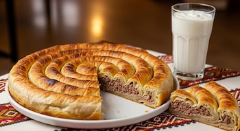 Burek bosnio en espiral, cortado para mostrar el relleno de carne picada, en un plato blanco. Un vaso de yogur y un mantel balcánico tradicional complementan la escena.