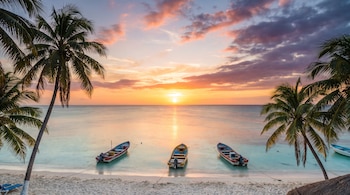 Playa de arena blanca con mar turquesa al atardecer, sol en el horizonte, reflejo dorado. Tres barcos de pesca en el agua, dos palmeras y nubes de colores.