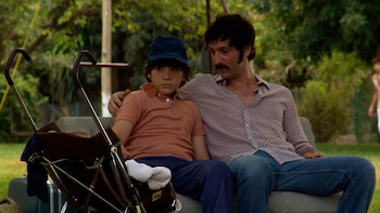 Un hombre con bigote y camisa estampada abraza a un niño con gorra azul y polo naranja, sentados en un banco con un cochecito al lado, en un entorno verde