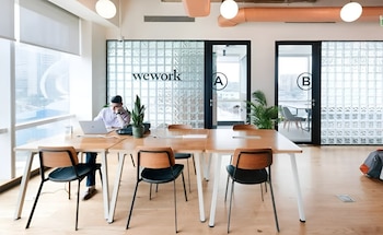 Hombre trabaja en laptop en una mesa de madera de oficina. Se ven varias sillas, plantas, grandes ventanas y paredes de bloques de vidrio con el logo WeWork