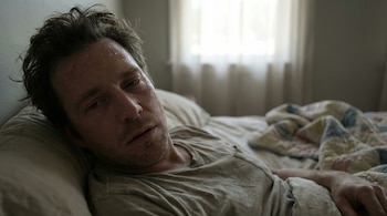 Primer plano de un hombre con pelo castaño revuelto y gotas de sudor en la frente, acostado en la cama con una camiseta gris, una ventana al fondo.