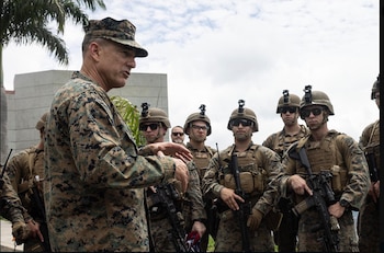Un oficial militar con uniforme de camuflaje habla con diez soldados equipados con cascos, gafas, chalecos tácticos y fusiles. Fondo de palmera y edificio