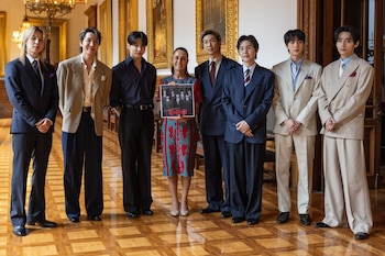 Una mujer con vestido estampado sostiene un cuadro con la imagen de BTS, mientras posa con los siete miembros del grupo K-pop en una sala elegante con piso de madera