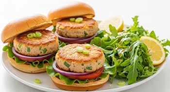 Tres hamburguesas de atún, papa y cebollita en pan, con lechuga, tomate y cebolla morada, en plato blanco, junto a ensalada verde con limón y un fondo claro.