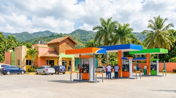 Gasolinera moderna y colorida con surtidores naranjas y verdes, varios autos y personas, un edificio de tonos vibrantes, palmeras y montañas al fondo bajo un cielo nublado.