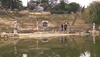Un estanque de agua refleja un anfiteatro de piedra con gradas, estatuas y dos personas en la base
