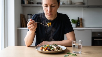 Mujer sentada, sosteniendo un tenedor con comida que emite vapor. Hay un plato con vegetales y granos, y un vaso de agua en la mesa de madera de una cocina.