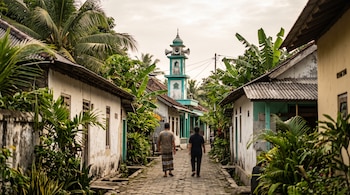 Dos personas caminan de espaldas por un camino de piedra flanqueado por casas blancas y plantas tropicales hacia un minarete verde.