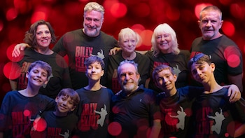 Un grupo de diez personas, incluyendo adultos y niños, sonríe a cámara. Todos llevan camisetas negras con el logo de 'Billy Elliot el Musical' sobre un fondo rojo con luces bokeh