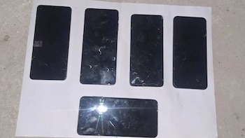 Cinco teléfonos móviles negros, cuatro en fila y uno debajo, sobre una hoja de papel blanco. Sus pantallas están oscuras y algunas con marcas de uso