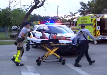 Dos paramédicos mueven a una persona en una camilla por la calle. Detrás se ven un SUV policial con luces intermitentes y una ambulancia de Miami-Dade Fire Rescue
