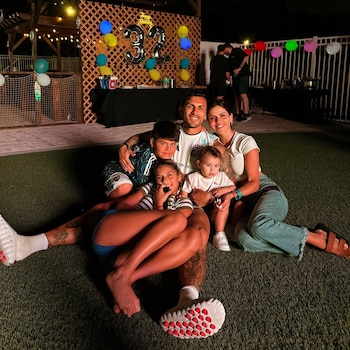 Cinco personas, un hombre, una mujer y tres niños, posan sentados en el césped. Hay globos de colores y el número 33 al fondo. Es de noche
